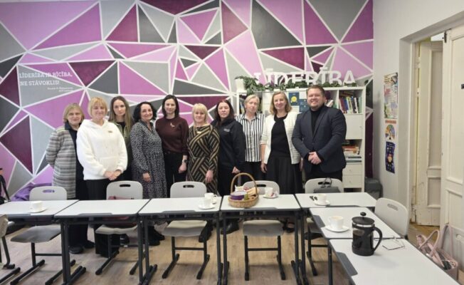 Vienpadsmit cilvēku (desmit sievietes un viens vīrietis) grupa stāv telpā ar ģeometrisku sienas gleznojumu violetos un pelēkos toņos. Uz sienas rakstīts: 