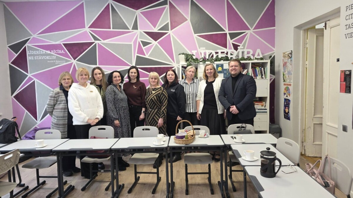 Vienpadsmit cilvēku (desmit sievietes un viens vīrietis) grupa stāv telpā ar ģeometrisku sienas gleznojumu violetos un pelēkos toņos. Uz sienas rakstīts: 