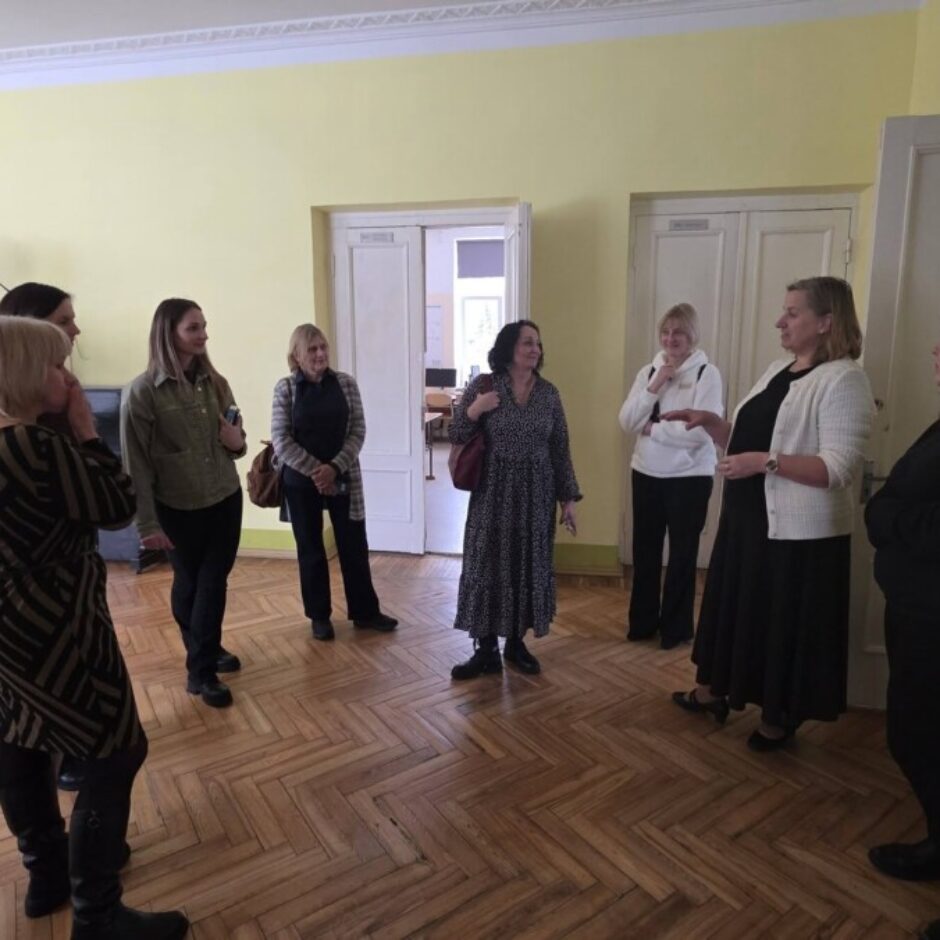 Grupas sieviešu sarunājas telpā ar dzeltenām sienām un koka parketa grīdu. Pie sienas redzama 