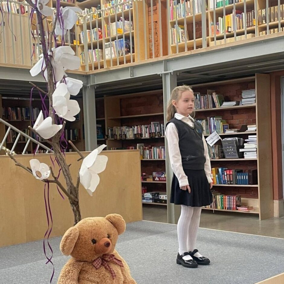 Jauna meitene skolas formā stāv uz nelielas skatuves, blakus sēž liels brūns plīša lācis uz koka celma pie dekoratīva koka ar baltiem papīra ziediem. Fonā redzami bibliotēkas grāmatu plaukti.