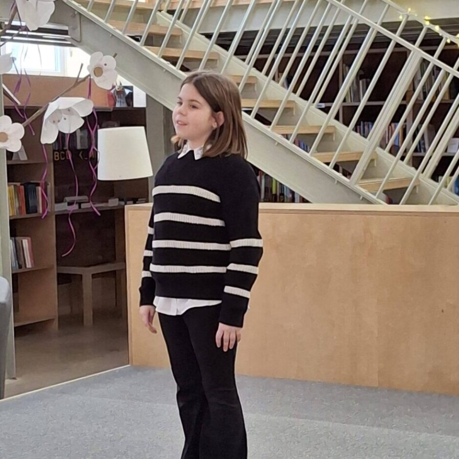 Jauna meitene melnā svītrainā džemperī un melnās biksēs stāv uz skatuves, Annija Meire (4.klase) skaita dzejoli Fonā ir grāmatu plaukti un metāla kāpnes.