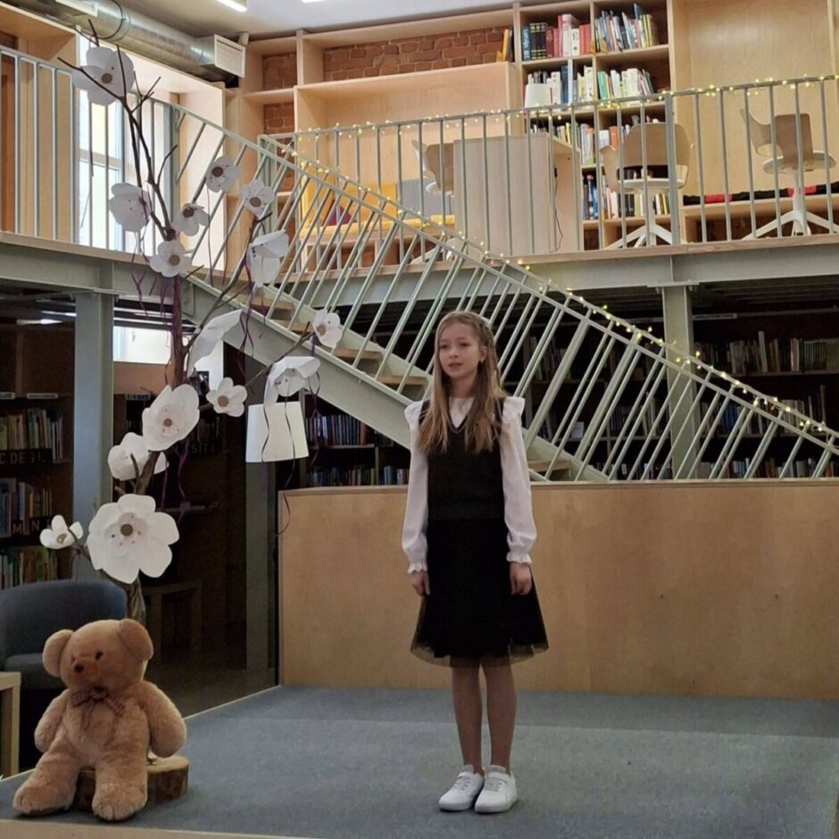 Jauna meitene stāv uz skatuves, Anete Roļskija (4.klase) skaita dzejoli Fonā ir grāmatu plaukti un metāla kāpnes.