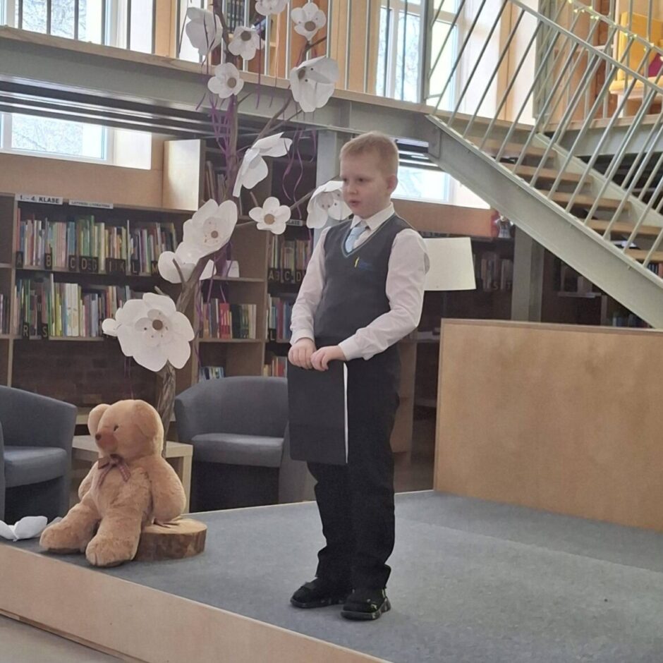 Stāv uz skatuves, Salvis Neļķe (1.a klase) skaita dzejoli Fonā ir grāmatu plaukti un metāla kāpnes.