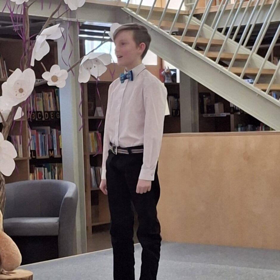 Stāv uz skatuves, Daniels Davidonis (4.klase) skaita dzejoli Fonā ir grāmatu plaukti un metāla kāpnes.