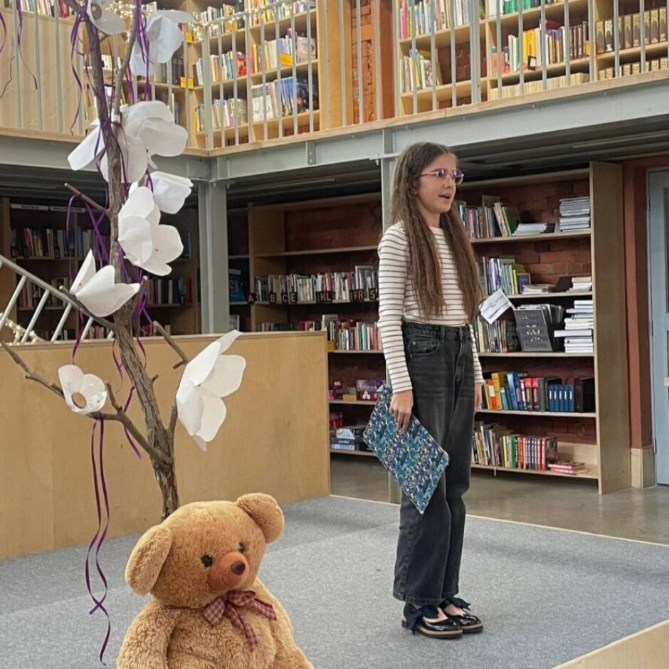 Jauna meitene stāv uz skatuves, Adrija Plūmane skaita dzejoli Fonā ir grāmatu plaukti un metāla kāpnes.