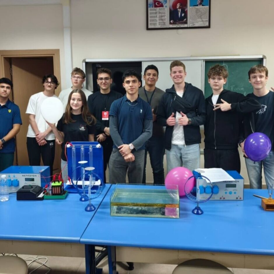 Vienpadsmit jauni cilvēki, viena meitene un desmit zēni, stāv aiz zila galda zinātnes klasē ar eksperimentu aprīkojumu, tostarp ierīci ar uzrakstu 