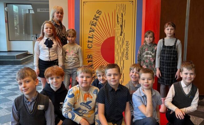 Bērnu grupa ar pieaugušo pozē uz sarkana paklāja, fonā milzīgs plakāts, kas izskatās pēc sērkociņu kastes. Uz plakāta redzams uzraksts 