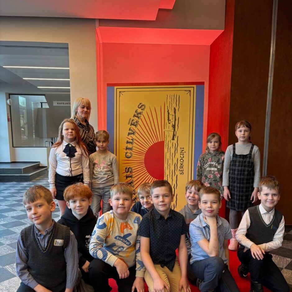 Bērnu grupa ar pieaugušo pozē uz sarkana paklāja, fonā milzīgs plakāts, kas izskatās pēc sērkociņu kastes. Uz plakāta redzams uzraksts 