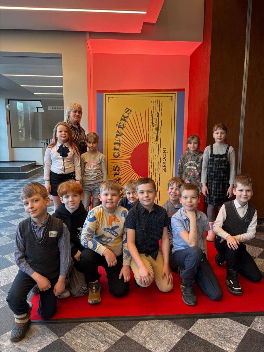 Bērnu grupa ar pieaugušo pozē uz sarkana paklāja, fonā milzīgs plakāts, kas izskatās pēc sērkociņu kastes. Uz plakāta redzams uzraksts 