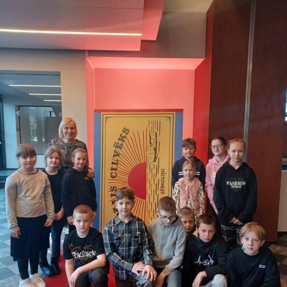 Grupa ar divpadsmit bērniem un vienu pieaugušo sievieti pozē, stāvot un tupot uz sarkana paklāja pie liela, zeltaini sarkana paneļa. Panelī ir vertikāls teksts 