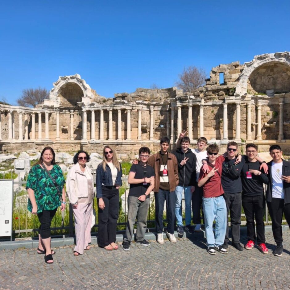 Grupa, kurā ir trīs sievietes un desmit jaunieši, pozē saulainā dienā pie liela seno drupas ar kolonnām un arkām.