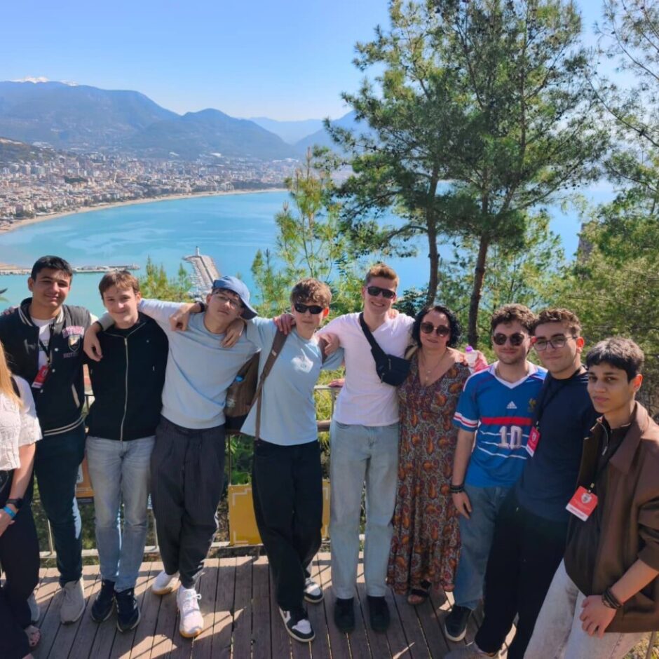 Desmit cilvēku grupa, tostarp jaunieši un pieaugušie, stāv uz koka platformas, pozējot fotogrāfijai. Aiz viņiem paveras plašs panorāmas skats uz piekrastes pilsētu ar ostu un kuģiem, lielu zilu līci un kalniem fonā. Apkārt redzami zaļi koki.