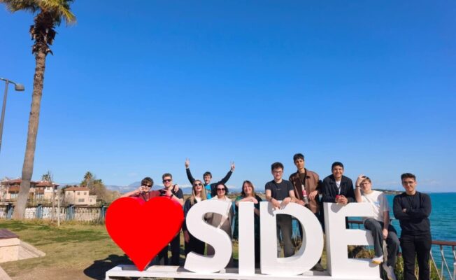 Jauniešu grupa pozē pie liela balta uzraksta, kas veido vārdu 