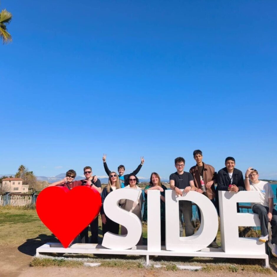 Jauniešu grupa pozē pie liela balta uzraksta, kas veido vārdu 