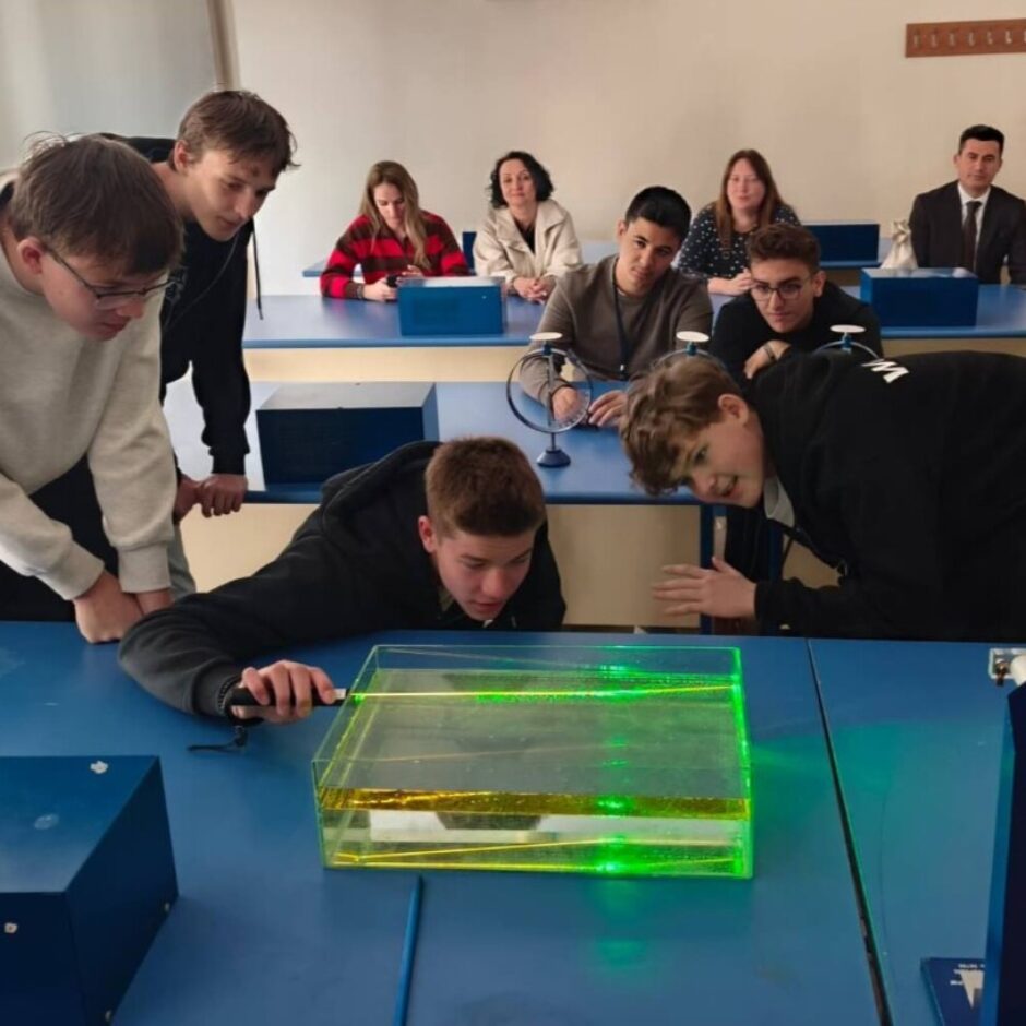 Skolēni fizikas klasē veic eksperimentu ar lāzeru. Priekšplānā trīs zēni noliecās pār caurspīdīgu tvertni ar šķidrumu, kurā redzami zaļi lāzera gaismas stari. Viens no zēniem tur lāzera rādītāju. Fonā redzami citi skolēni un pieaugušais.