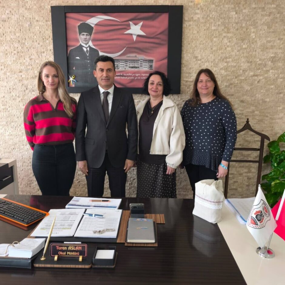 Četri cilvēki, vīrietis un trīs sievietes, stāv pie liela biroja galda. Uz galda ir vārda plāksnīte ar uzrakstu 