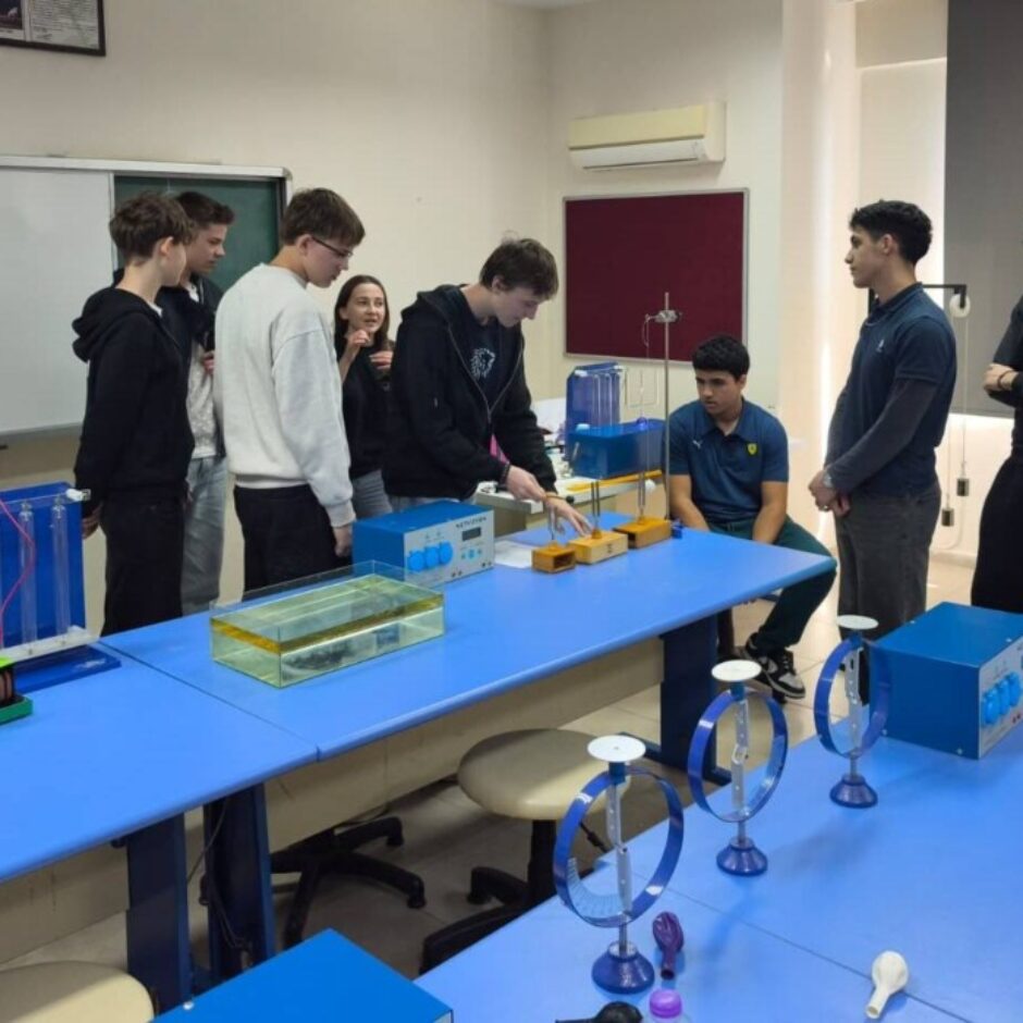 Grupa studentu stāv ap ziliem galdiem zinātnes laboratorijā vai klasē, kur notiek eksperiments. Viens students aktīvi darbojas ar ierīcēm, ko ieskauj citi vērojoši studenti.