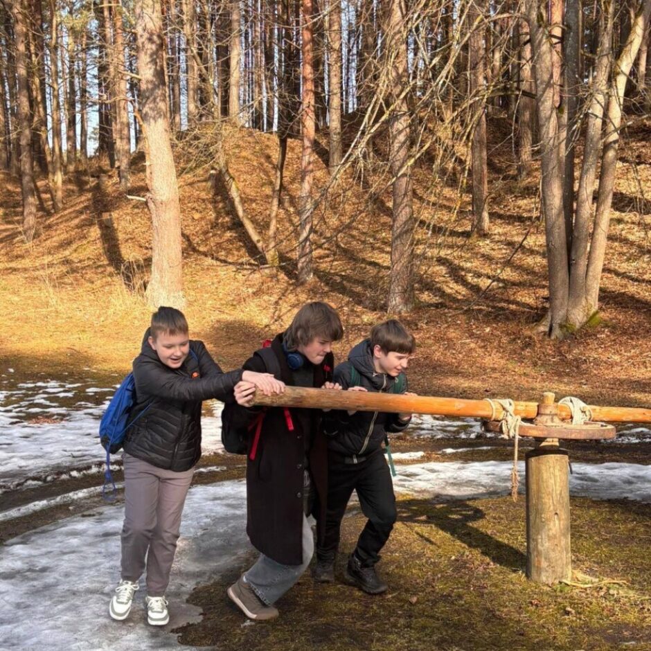 Trīs zēni darbojas ar lielu koka sviru āra ekspozīcijā, fonā kaili koki un uz zemes sniega laukumi.