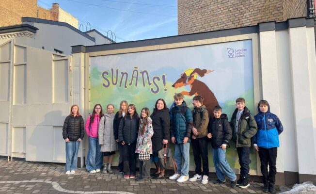 Grupa bērnu un viens pieaugušais stāv ārā, pozējot pie ēkas sienas, uz kuras ir liels plakāts. Uz plakāta redzams skrejošs brūns suns ar dzeltenu lakatiņu ap kaklu, un teksts 