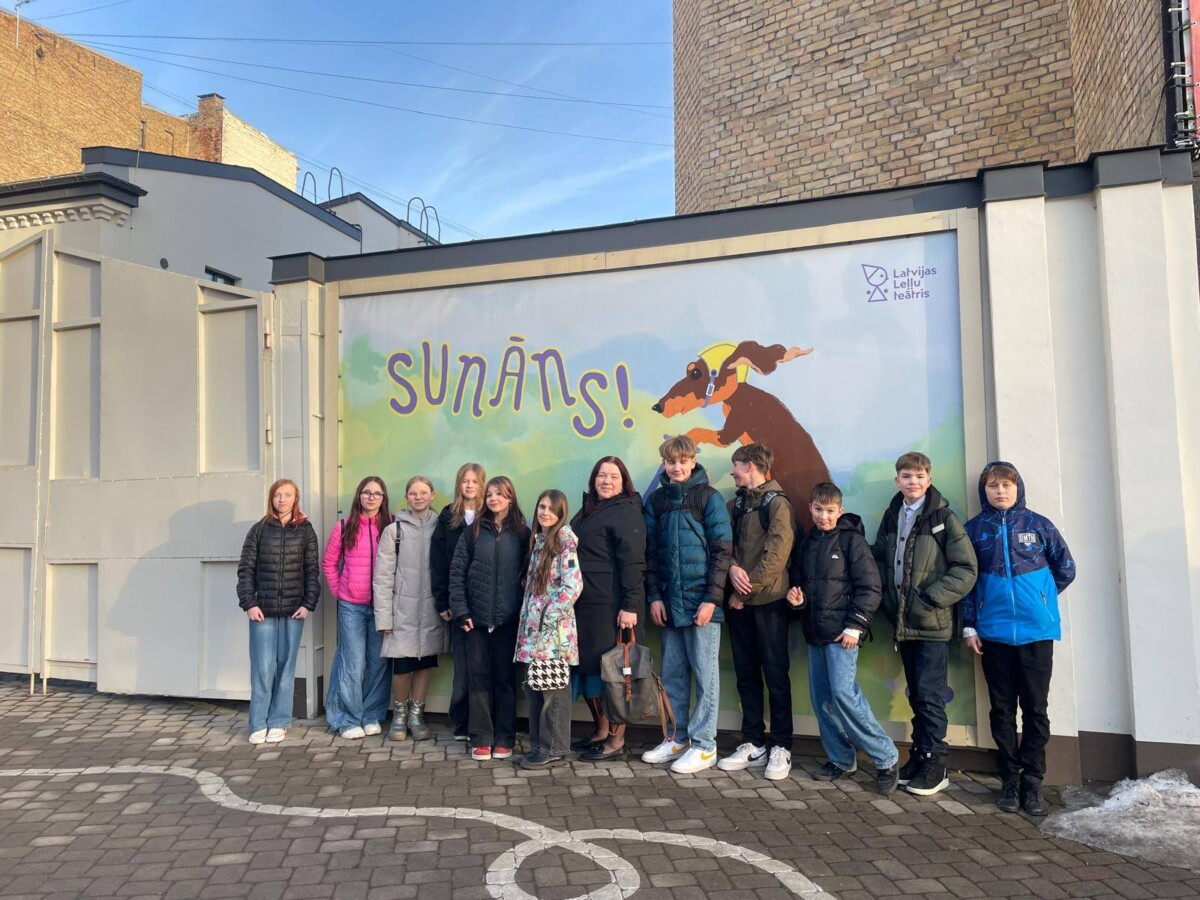 Grupa bērnu un viens pieaugušais stāv ārā, pozējot pie ēkas sienas, uz kuras ir liels plakāts. Uz plakāta redzams skrejošs brūns suns ar dzeltenu lakatiņu ap kaklu, un teksts 
