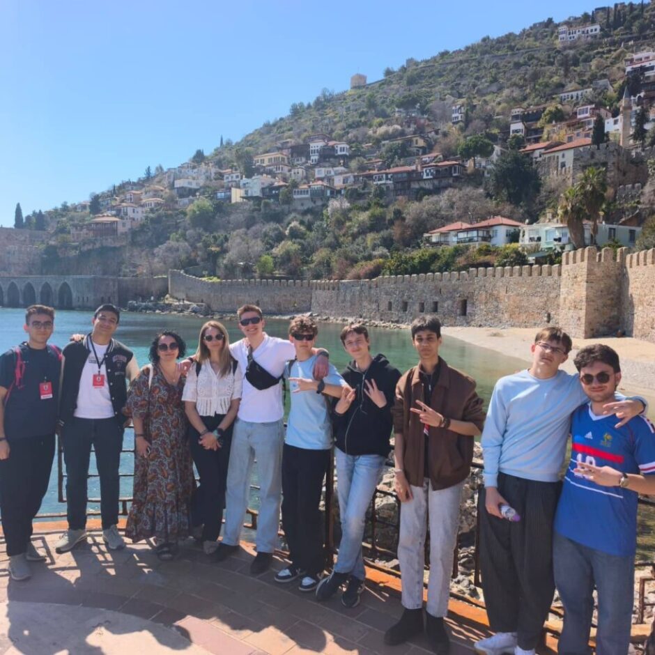 Desmit jaunieši stāv pie dzelzs margām, fonā jūra, seni akmens mūri un daudzas mājas uz stāva kalna.