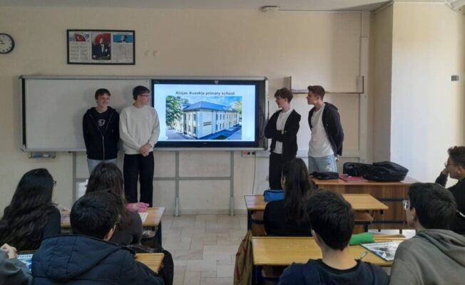 Četri jaunieši prezentē klasē pie interaktīvā ekrāna, uz kura redzama ēka un teksts 