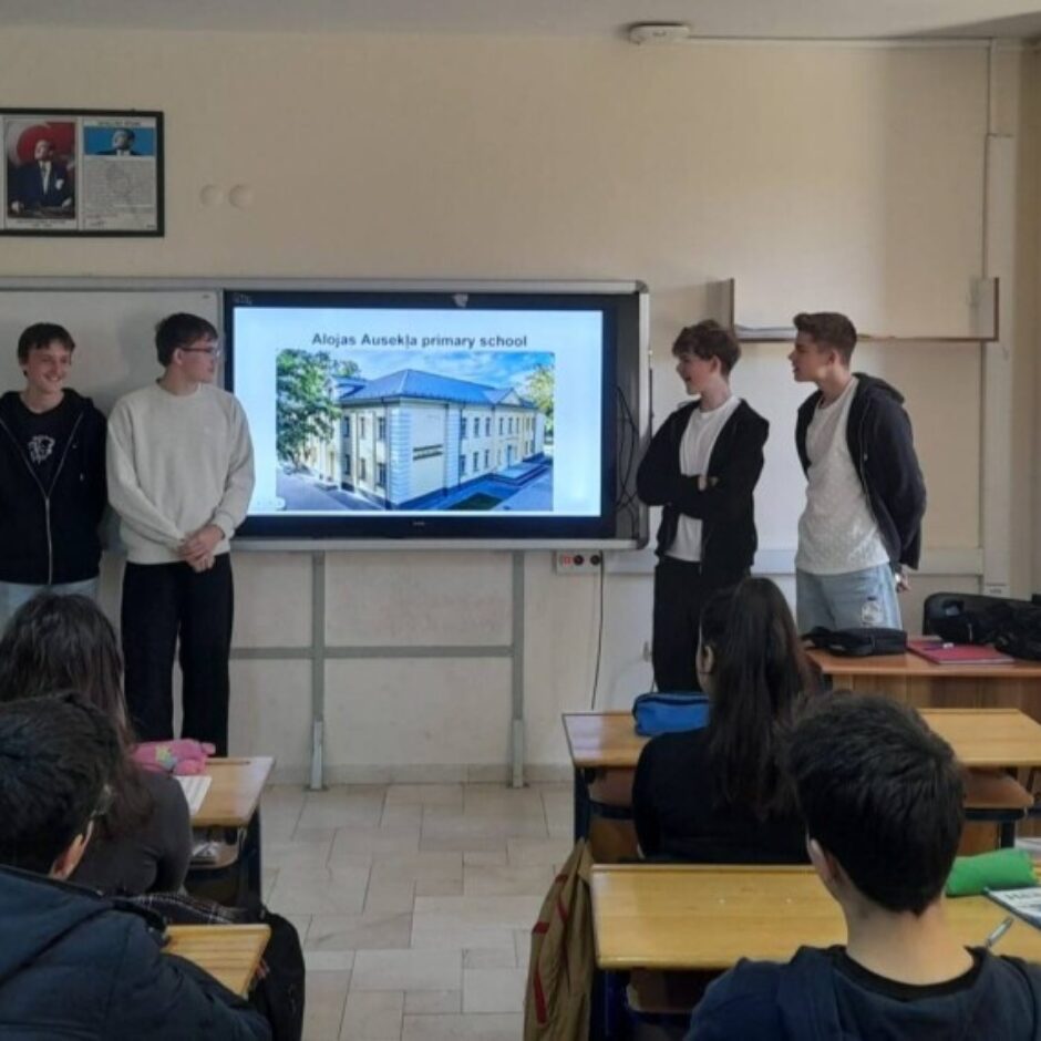 Četri jaunieši prezentē klasē pie interaktīvā ekrāna, uz kura redzama ēka un teksts 