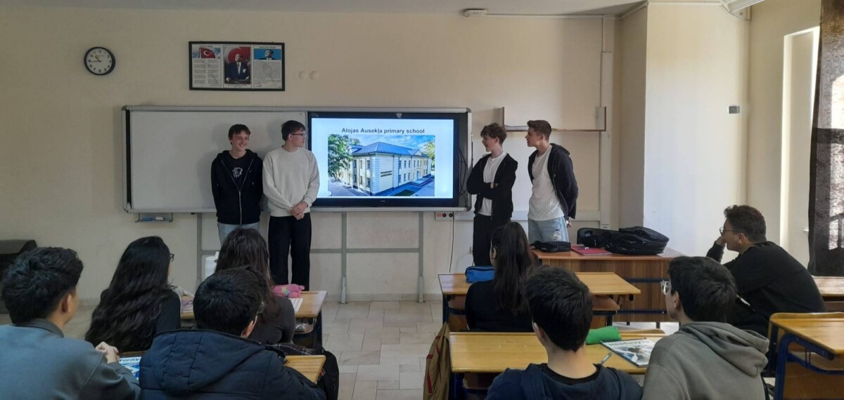 Četri jaunieši prezentē klasē pie interaktīvā ekrāna, uz kura redzama ēka un teksts 