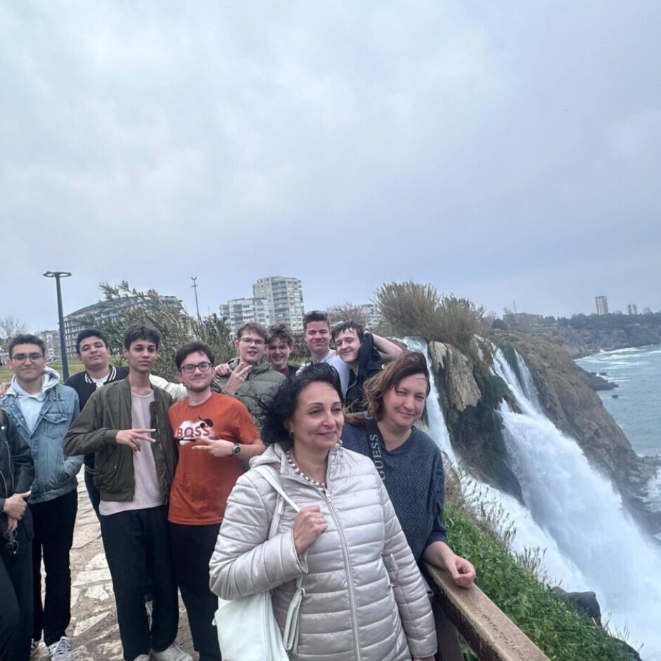 Smaidīga cilvēku grupa, galvenokārt jauni pieaugušie un pusaudži, pozē blakus lielam ūdenskritumam, kas gāžas jūrā. Priekšplānā divas sievietes smaida, un aiz viņām rinda jauniešu skatās kamerā. Jūra stiepjas pa labi, un tālumā zem apmākušām debesīm redzamas pilsētas ēkas.