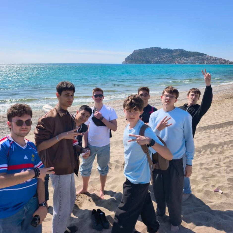 Jaunieši pozē saulainā smilšu pludmalē pie zilas jūras. Daži rāda miera zīmes. Fonā redzams liels, zaļš kalns, kas paceļas no jūras.