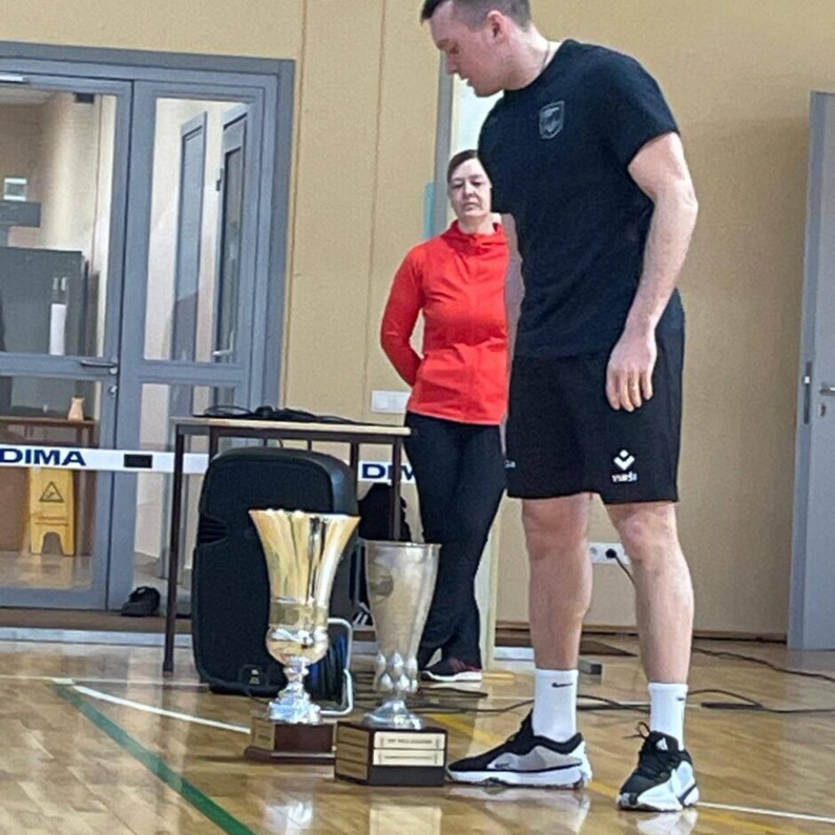 Garš vīrietis melnā sporta tērpā stāv sporta zālē, skatoties uz divām trofejām uz koka grīdas. Aiz viņa redzama sieviete sarkanā jakā