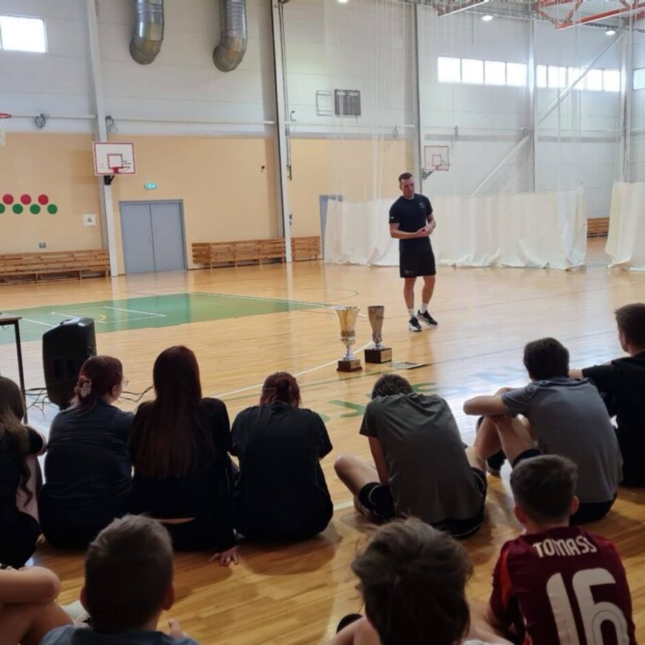 Sporta zālē vīrietis melnā apģērbā stāv uz koka grīdas un runā ar jauniešu grupu, kas sēž priekšā. Blakus vīrietim uz grīdas stāv divi zelta kausi. Kreisajā pusē sieviete sarkanā kreklā stāv pie staba.