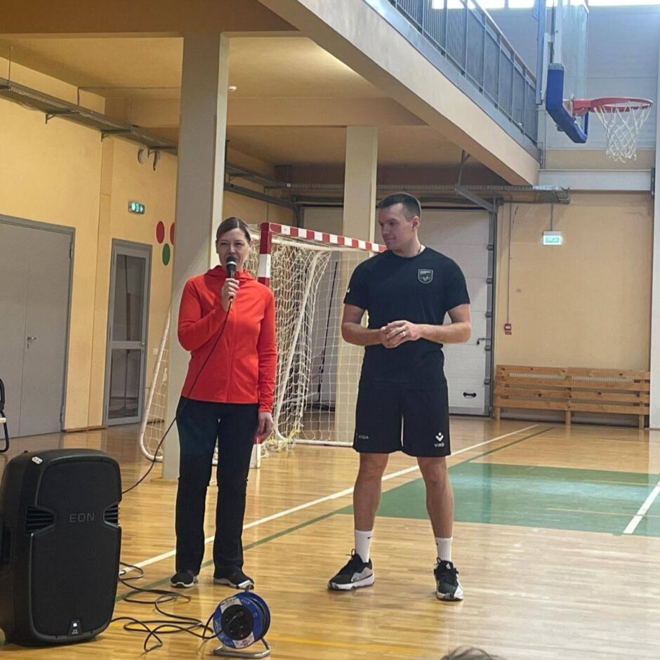 Sieviete sarkanā sporta jakā runā mikrofonā, stāvot sporta zālē blakus vīrietim melnā sporta tērpā ar logotipu uz T-krekla un šortiem ar uzrakstu 