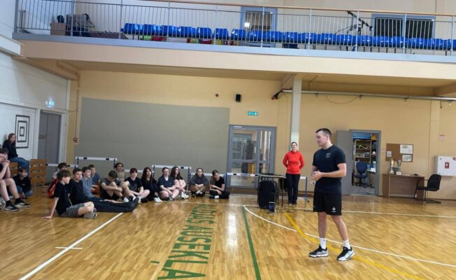 Vīrietis melnā sporta tērpā stāv sporta zālē un runā ar pusaudžu grupu, kas sēž uz koka grīdas. Fonā stāv sieviete sarkanā kreklā.