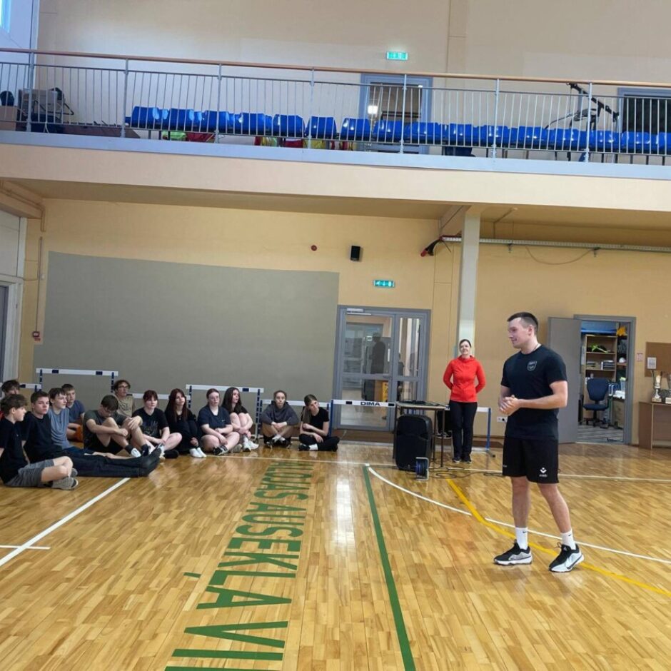 Vīrietis melnā sporta tērpā stāv sporta zālē un runā ar pusaudžu grupu, kas sēž uz koka grīdas. Fonā stāv sieviete sarkanā kreklā.