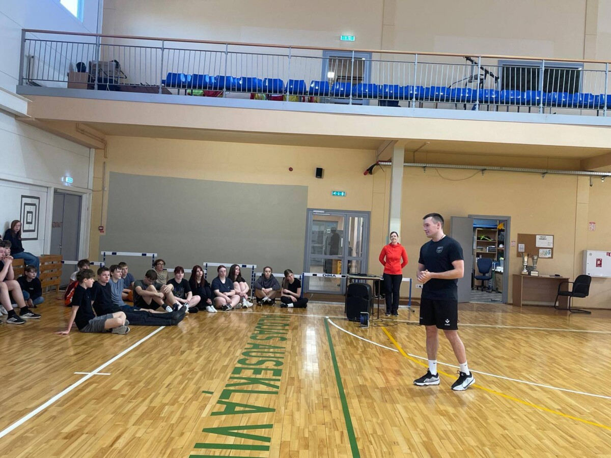 Vīrietis melnā sporta tērpā stāv sporta zālē un runā ar pusaudžu grupu, kas sēž uz koka grīdas. Fonā stāv sieviete sarkanā kreklā.