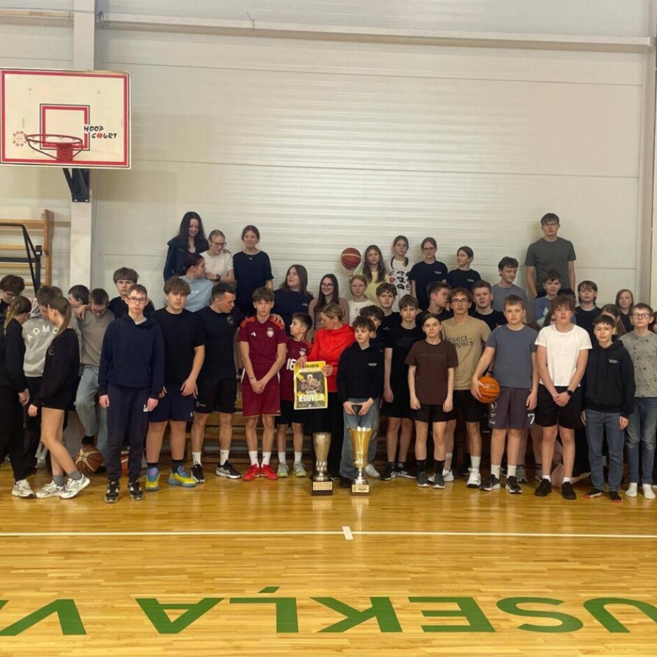 Liela jauniešu un pieaugušo grupa pozē grupveida foto basketbola zālē. Priekšplānā uz grīdas ir divi zelta kausi un basketbola bumba.
