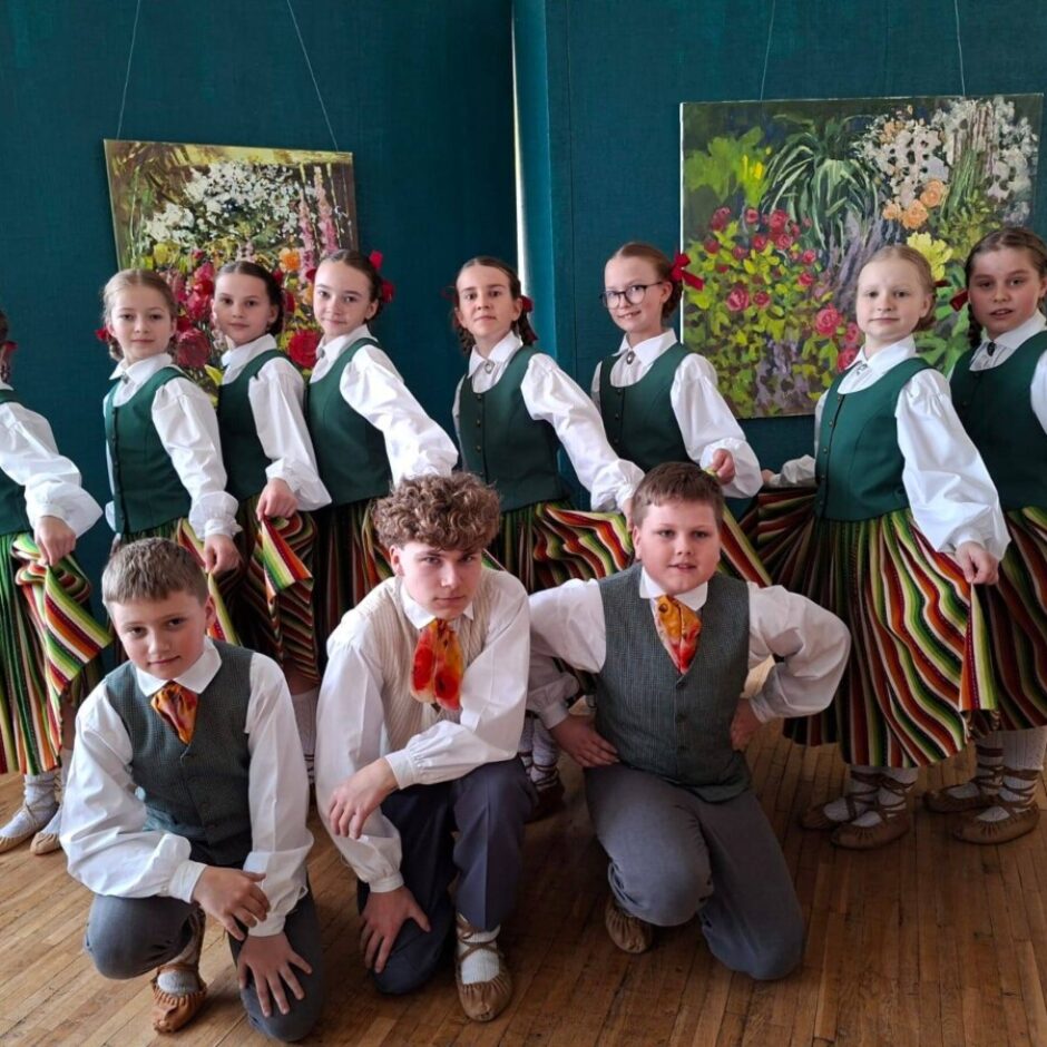 Bērnu grupa latviešu tautas tērpos pozē fotogrāfijai uz koka grīdas, zilas sienas un divu ziedu gleznu fonā. Aizmugurē stāv deviņas meitenes baltās blūzēs, zaļās vestēs un svītrainos svārkos, priekšplānā tup trīs zēni baltos kreklos un vestēs.