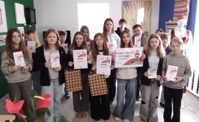 Grupa skolēnu, meitenes un zēni, stāv telpās un lepni demonstrē saņemtos diplomus, atzinības rakstus un dāvanu maisiņus. Daži no diplomiem vēsta 