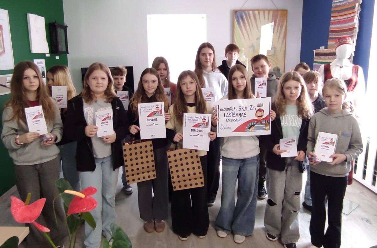 Grupa skolēnu, meitenes un zēni, stāv telpās un lepni demonstrē saņemtos diplomus, atzinības rakstus un dāvanu maisiņus. Daži no diplomiem vēsta 