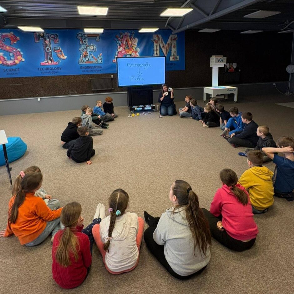 Bērni sēž aplī uz grīdas un vēro pasniedzēju, kura demonstrē ar maziem objektiem. Aiz viņas ekrānā redzams teksts 