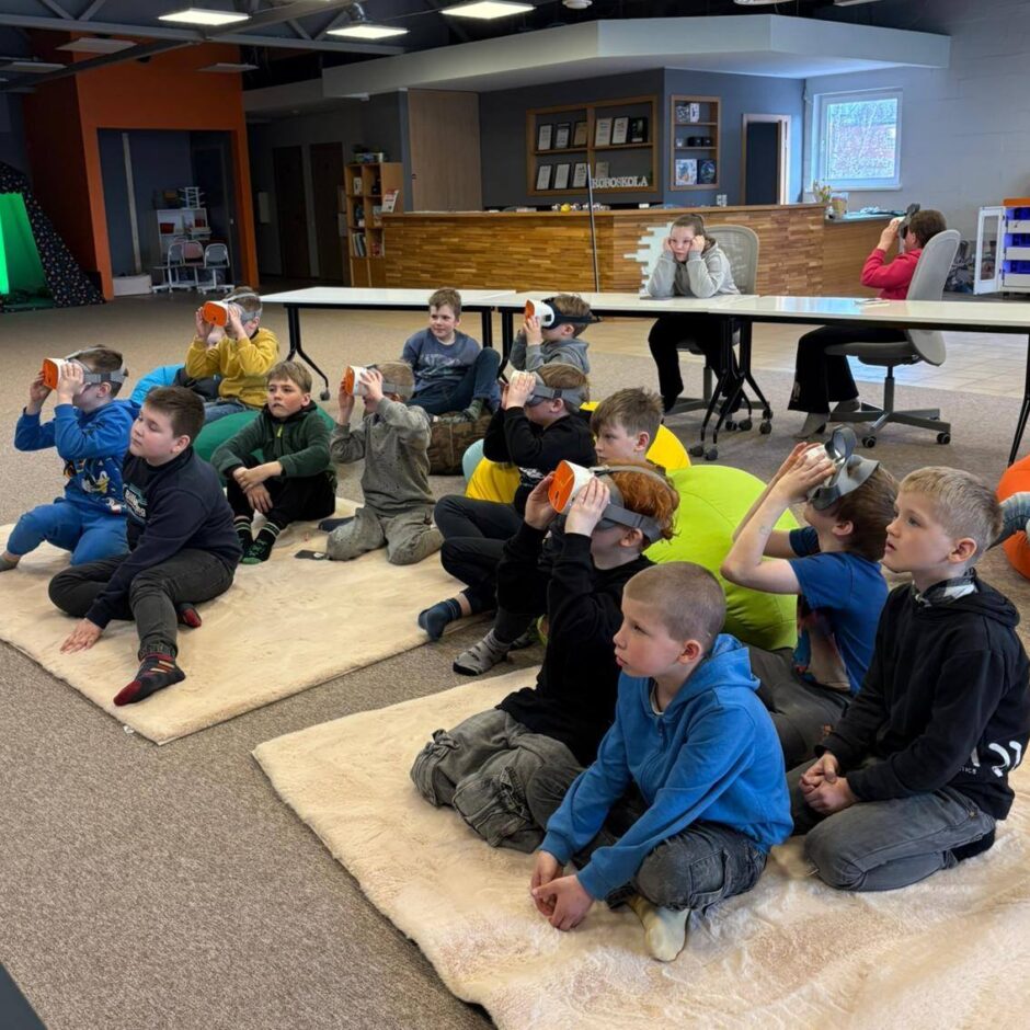 Duci bērnu, galvenokārt zēni, sēž uz grīdas uz paklājiem un pufiem, valkājot virtuālās realitātes brilles un vērojot saturu. Fonā redzama moderna telpa ar izkārtni ROBOSKOLA.