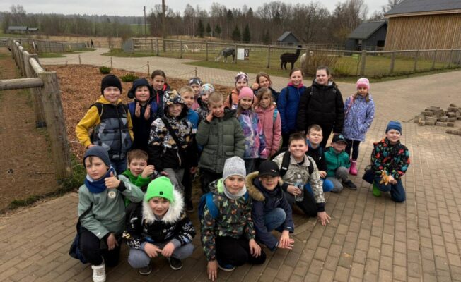 Liela bērnu grupa pozē fotogrāfijai alpakas fermā. Viņi stāv un tup uz bruģēta laukuma, fonā redzamas alpakas iežogojumos un koka ēkas.