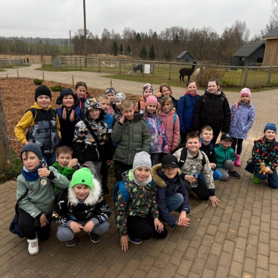 Liela bērnu grupa pozē fotogrāfijai alpakas fermā. Viņi stāv un tup uz bruģēta laukuma, fonā redzamas alpakas iežogojumos un koka ēkas.