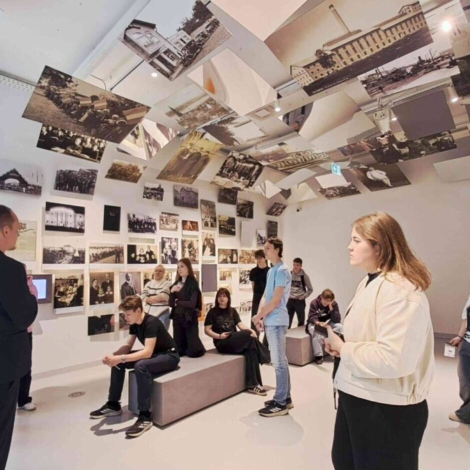 Muzeja izstādes telpa, kur jauniešu grupa klausās gida stāstījumā. Sienas un griesti ir klāti ar daudzām melnbaltām vēsturiskām fotogrāfijām. Kreisajā pusē uz balta paneļa redzams teksts 