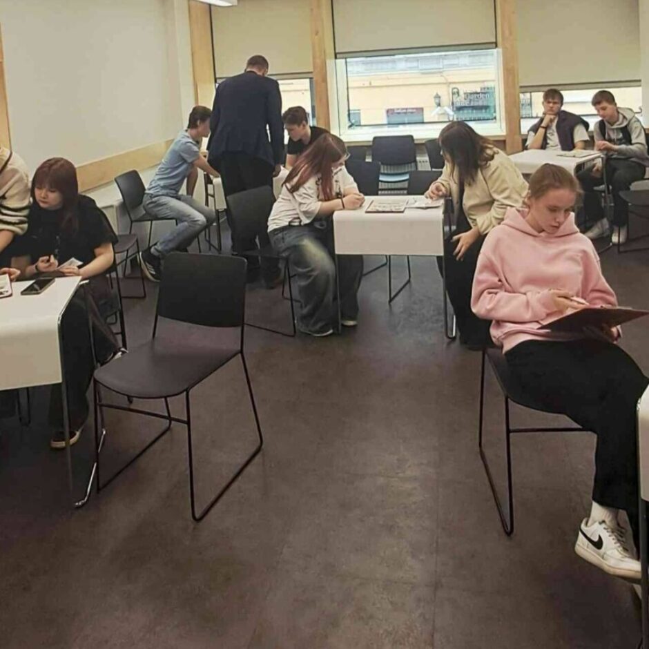 Vairāki jaunieši sēž pie baltiem galdiem gaišā telpā. Priekšplānā trīs meitenes ir pie galda, viena sieviete rāda uz materiāliem uz galda. Cita meitene raksta uz mapes, kamēr blakus esošā meitene valkā pelēku džemperi ar uzrakstu 