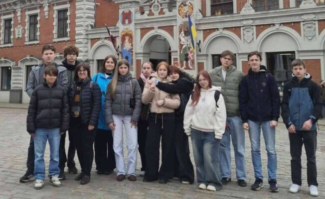 Grupa pusaudžu un pieaugušo pozē fotogrāfijai Rīgas Rātslaukumā, Melngalvju nama priekšā. Ornatā sarkanu ķieģeļu ēka ar baltiem rotājumiem un tornīšiem ir galvenais fons. Uz tās fasādes ir astronomiskais pulkstenis ar uzrakstiem 