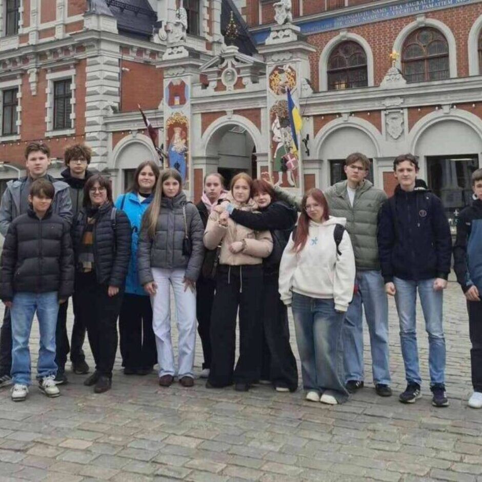 Grupa pusaudžu un pieaugušo pozē fotogrāfijai Rīgas Rātslaukumā, Melngalvju nama priekšā. Ornatā sarkanu ķieģeļu ēka ar baltiem rotājumiem un tornīšiem ir galvenais fons. Uz tās fasādes ir astronomiskais pulkstenis ar uzrakstiem 