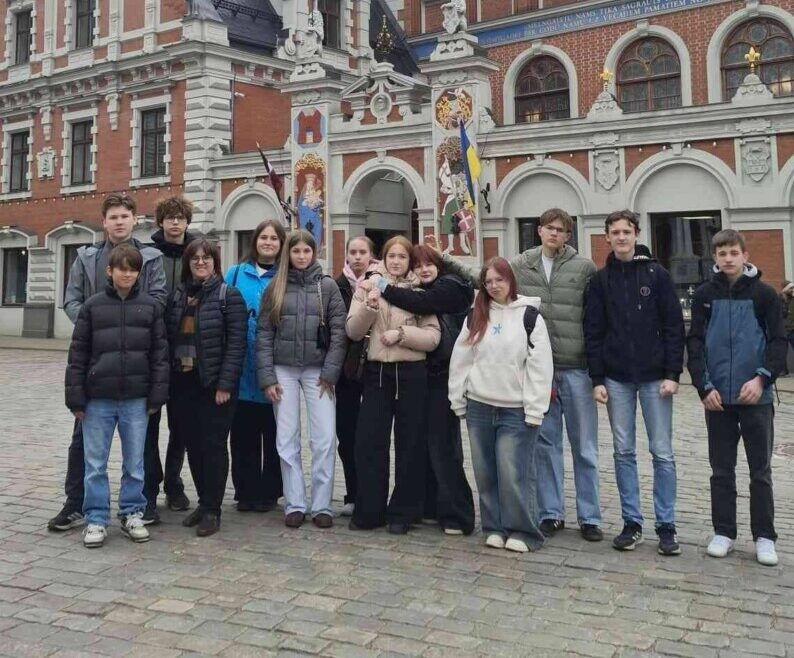 Grupa pusaudžu un pieaugušo pozē fotogrāfijai Rīgas Rātslaukumā, Melngalvju nama priekšā. Ornatā sarkanu ķieģeļu ēka ar baltiem rotājumiem un tornīšiem ir galvenais fons. Uz tās fasādes ir astronomiskais pulkstenis ar uzrakstiem 