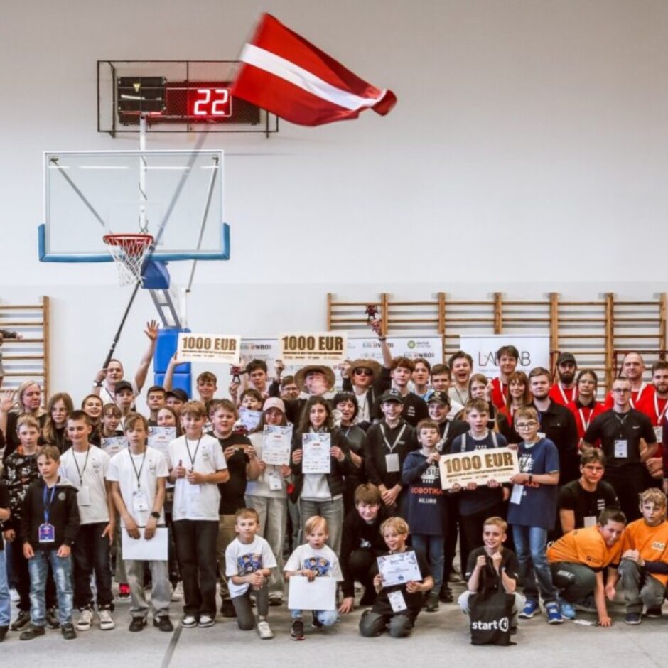 Liela bērnu, pusaudžu un pieaugušo grupa pozē sporta zālē, visticamāk, apbalvošanas ceremonijā. Daudzi jaunieši tur rokās sertifikātus un balvas, tostarp zīmes ar tekstu 